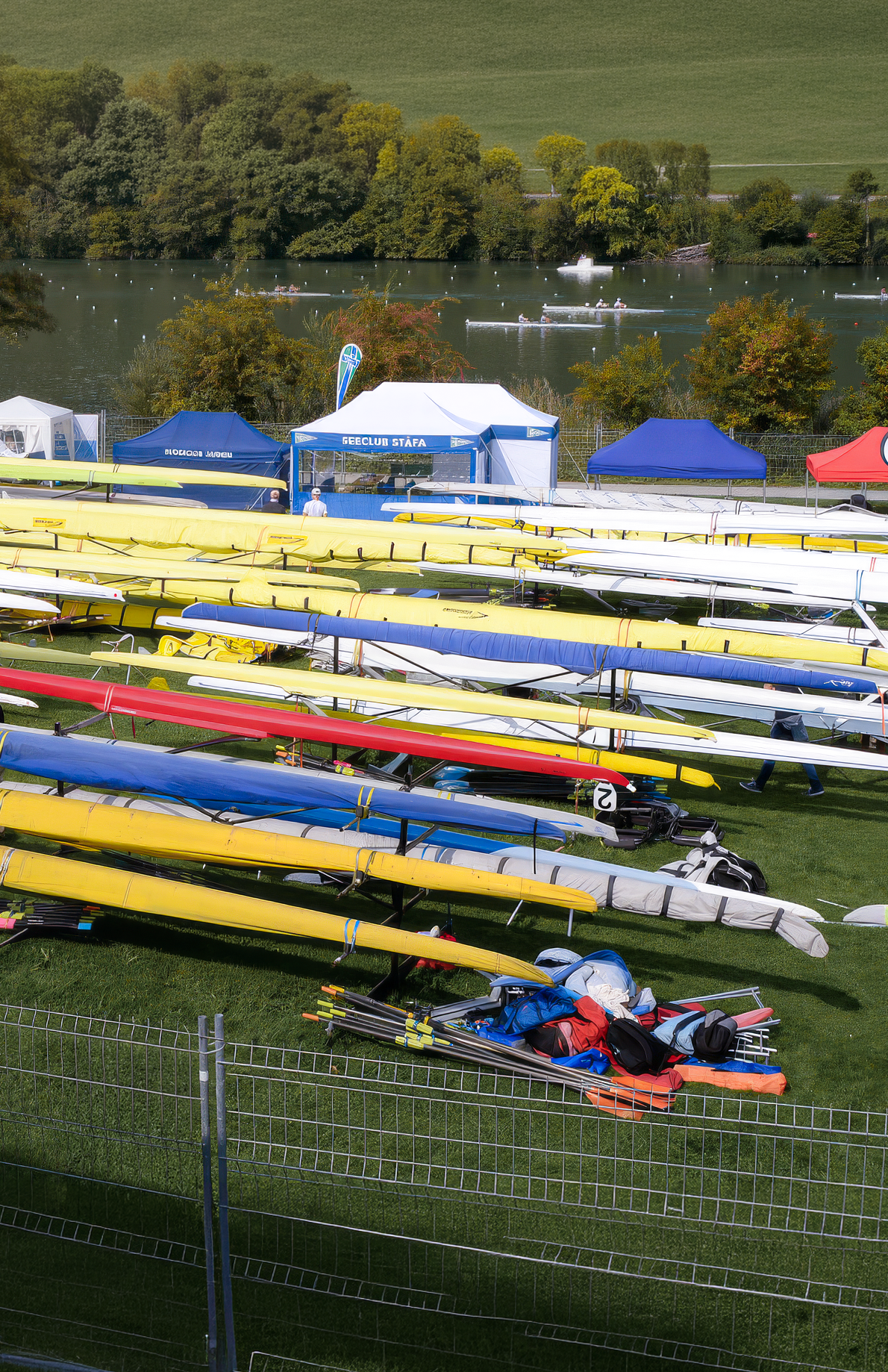 Image du lac du Rotsee dans le canton de Lucerne lors des championnats suisses d'aviron, on y voit au premier plan le parc à bateau, avec de multiple embarcations et, plus loin, les bateaux en pleine régate.